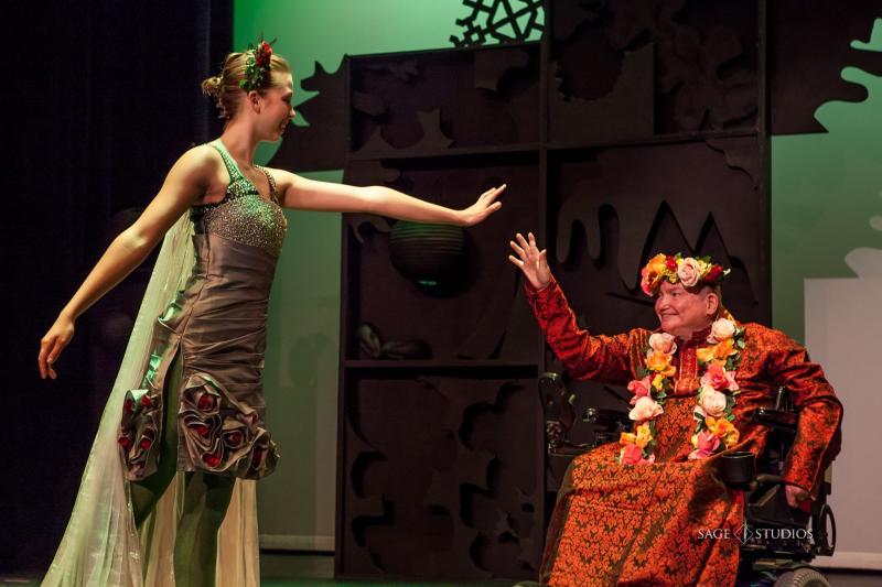 An actress wearing a green costume (adorned with decorative flowers) reaches for another actor that is in a wheelchair and wearing an orange and brown costume with a floral necklace and headdress.