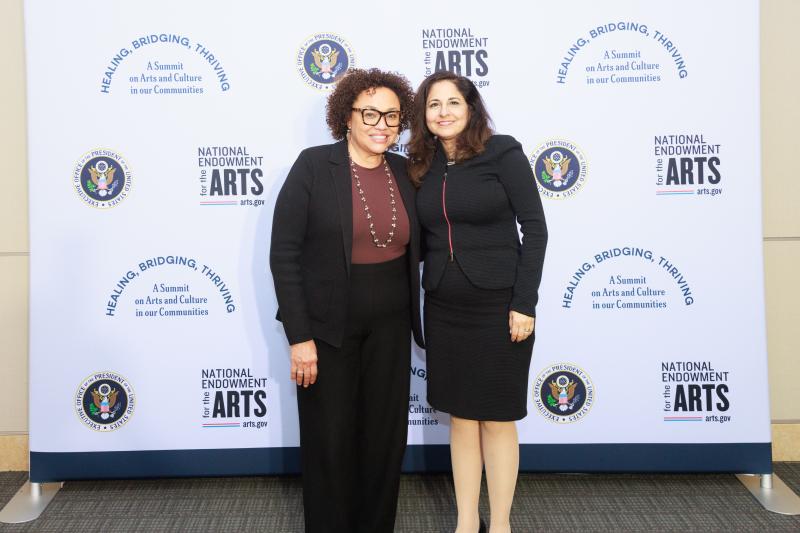 Woman with black curly hair wearing glasses and wearing a suite standing next to woman with long straight black hair wearing a dress.