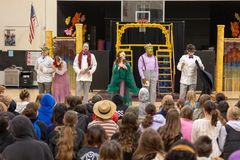 Six actors in elaborate colorful masks perform a Shakespeare play in a school gym
