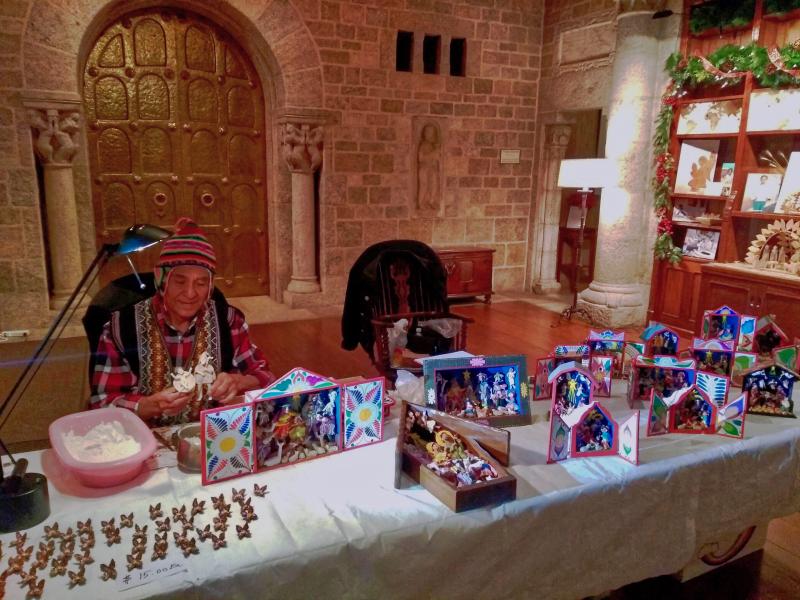 Jeronimo Lozano Retablo Great Hall Demonstration. Photo courtesy of © Glencairn Museum, Bryn Athyn, Pennsylvania A man sits with many colorful, intricate, hand-painted Nativities displayed on a table in front of him.