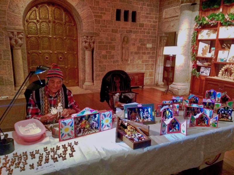 A man sits with many colorful, intricate, hand-painted Nativities displayed on a table in front of him.