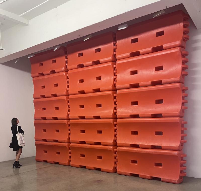 A woman stands inside a room where one wall is taken up by a stack of large red-orange plastic interlocking blocks.