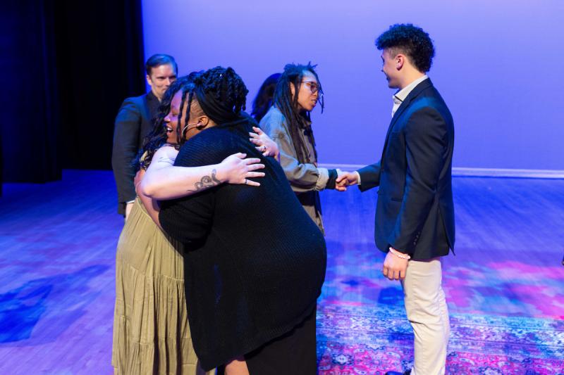 A group of people stand on stage, two people shake hands in the background and two people hug in the foreground.