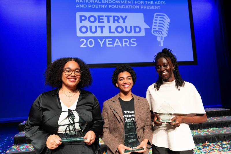 Three students smile in front of Poetry Out Loud backdrop holding trophies.