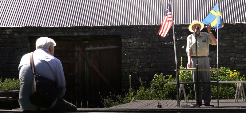 A ranger standing between two flapgs in front of a long low-slung building giving a presentation to an older man