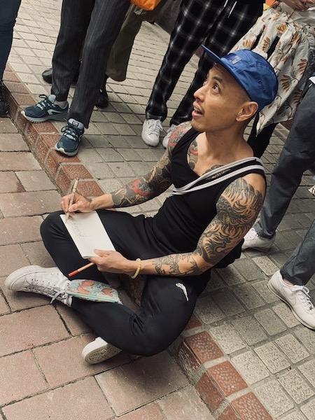 Marcos Chin, a thin Asian-Canadian man sits cross-legged on a sidewalk drawing
