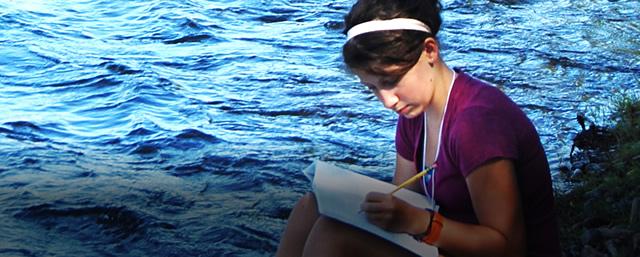 Young woman sitting by the waters edge writing in her notebook
