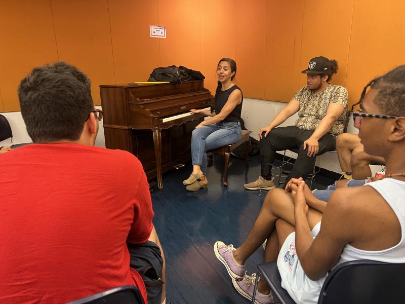 Four actors sit in chairs as a woman (center) plays on a piano