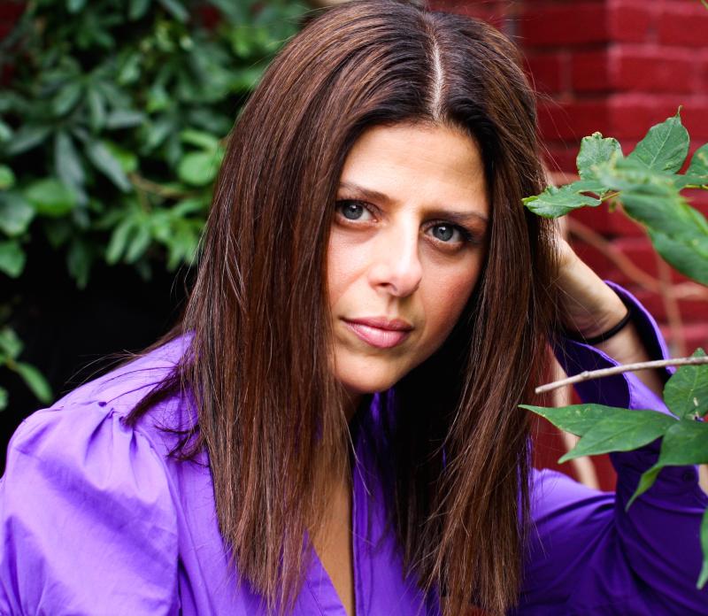 Nathalie Handal, who has brown hair and green eyes, smiles at the camera, wearing a purple shirt.