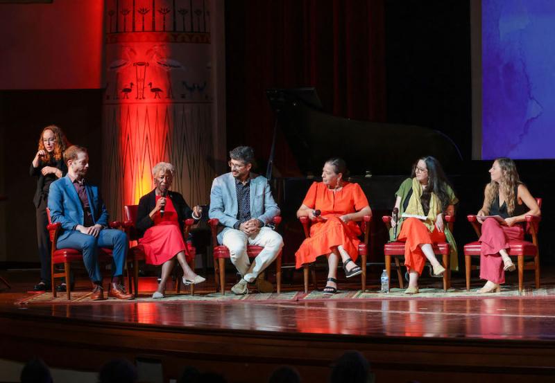 Six people sit in chairs on a stage looking at one of them speaking into a microphone. Behind them is a sign language interpreter