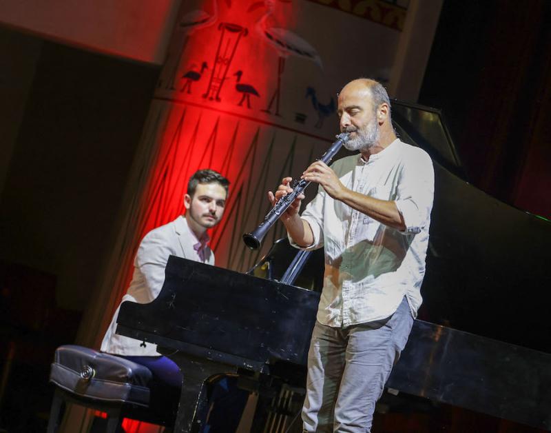 A young man sits at a piano looking at another man standing in front of the piano performing on the clarinet