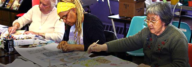 Three senior women at a long table covered with art supplies painting and drawing