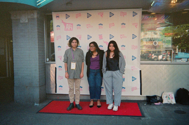 A Native man (left) and two women (center and right) stand in front of a film festival backdrop (with pink and blue film festival logos) and red step-and-repeat carpet.