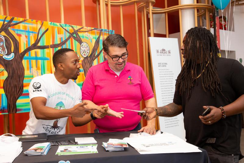 Two men in white and pink shirts hand a pamphlet to a man in a black shirt.