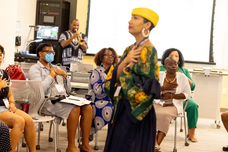 Black woman in yellow hat and flowery shirt stands in foreground with hand on her heart, group of people seated behind her also have hands over their hearts, and Black man at back plays a trumpet. 