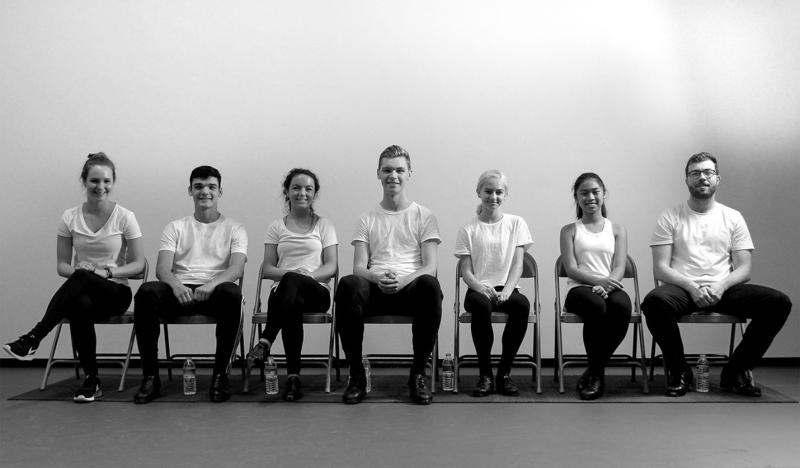 Black and white photo of seven dancers sitting in chairs and smiling.