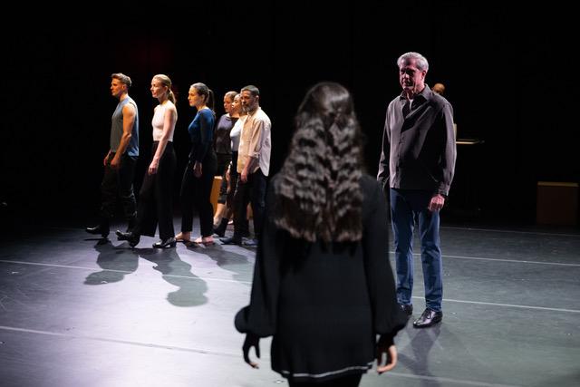Nine dancers on a stage (seven of the dancers looking outward toward the audience, one female cast member with her back facing the audience, and one male cast member standing upstage).