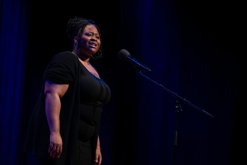 Young Black woman in black dress on stage speaking into a microphone.