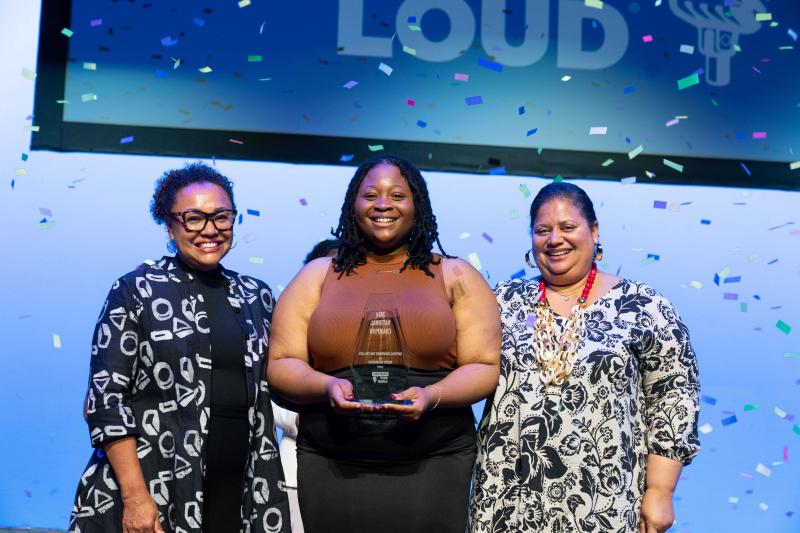 Three women stand smiling on a stage as confetti fall around them, the young woman in center holds a trophy