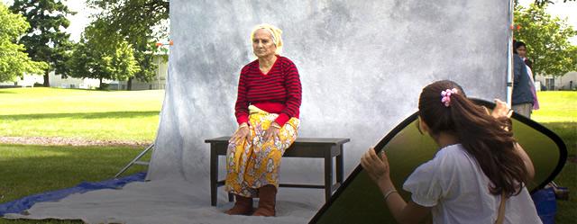 Older woman sitting outdoors on a bench in front of a large stone sculpture with a young person looking on.