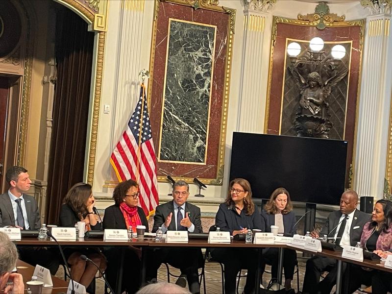 A group of people around a table look towards a person in the center speaking. Behind them is an American flag.