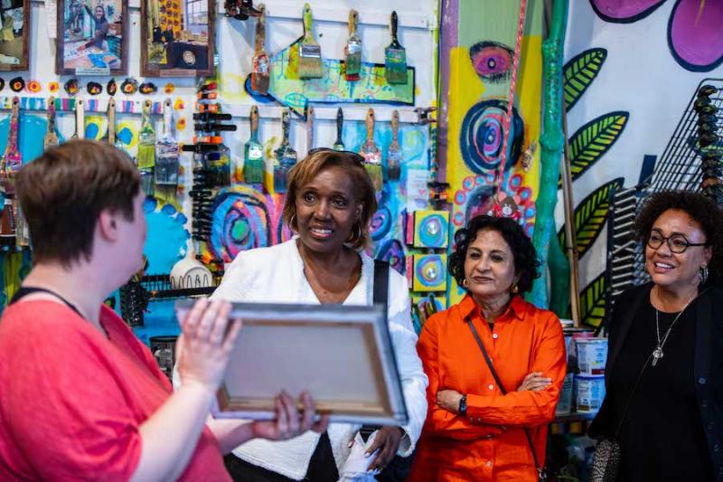 An individual with their back to the camera holds up a piece of art for three women to see.