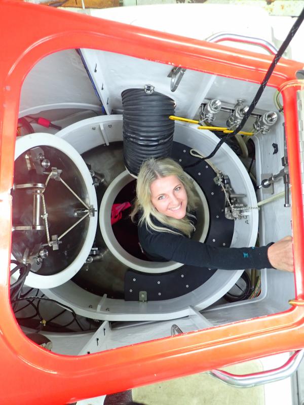White woman wearing black longsleeved shirt, emerging from a submersible that has a red exterior lining. White woman wearing black longsleeved shirt, emerging from a submersible that has a red exterior lining.
