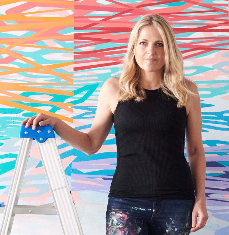 White woman in a black tank top and denim jeans holding on to a ladder, in front of a backdrop that has purple/orange/blue/green/red splatter paint. White woman in a black tank top and denim jeans holding on to a ladder, in front of a backdrop that has purple/orange/blue/green/red splatter paint.