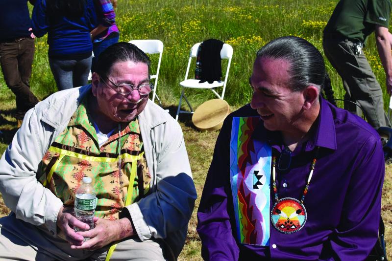 Elderly man wearing glasses and holding a bottle of water talking with a younger man next to him, smiling.