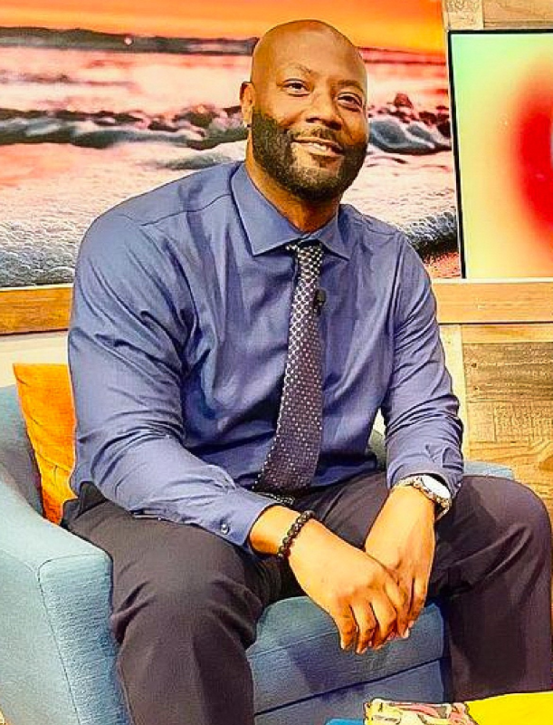 a middle-aged Black man wearing a shirt and tie sits on a chair smiling at the camera
