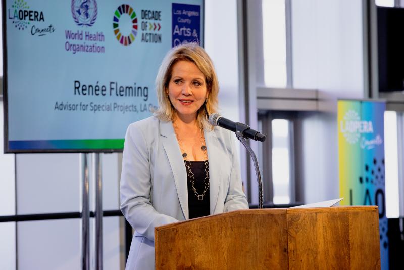 Woman with short light brown hair wearing a light blue blazer standing at a lectern.