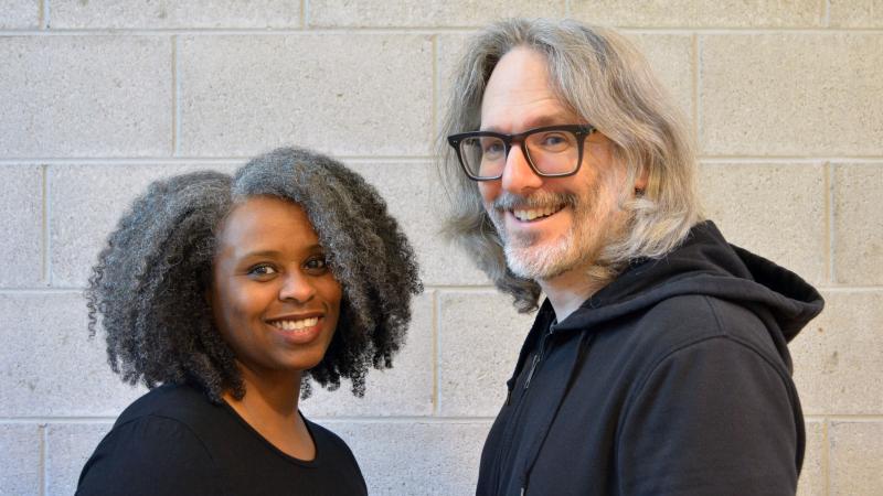 photo of a Black woman with long curly hair and a white man with longish hair, beard and glasses with a cinderblock wall in the background