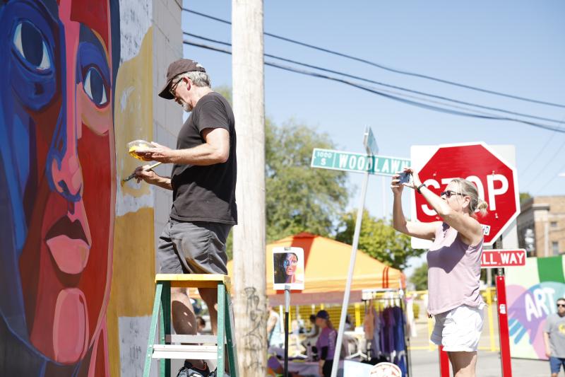 A man paints a mural on the side of a building while a woman takes a photo with her cell phone.