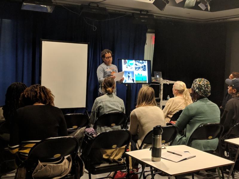 At a scriptwriting workshop in Spring 2020, a group of students sits in folding chairs facing an instructor reading from a sheet of paper.