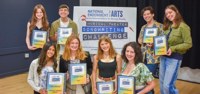 A group of teenagers smile and pose in front of a sign for the Musical Theater Songwriting Challenge. They each hold a binder with their name and the name of their winning song.