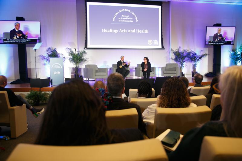 Asian man and Black/Latina woman on stage with screen behind them, audience members in chairs facing them. 
