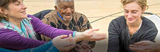 An older women, an older man, and a young woman in chairs in a gym holding hands out to the center 