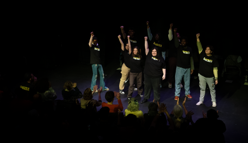Eight actors onstage (facing an audience) in black shirts with their hands up in the air (making a fist)