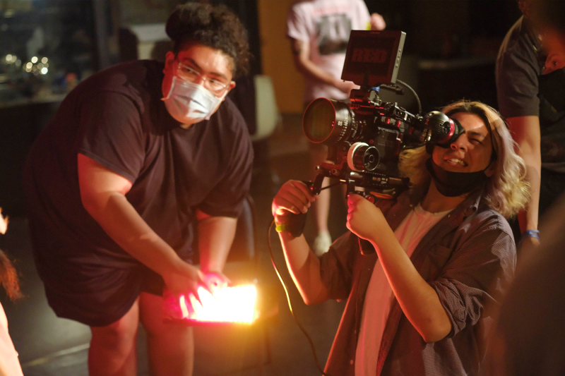 On a film set: Native filmmaker (left) wears a black t-shirt an and has a white mask on his face. Native filmmaker (right) wears a black shirt and has a black mask hanging on his chin while holding up camera equipment.