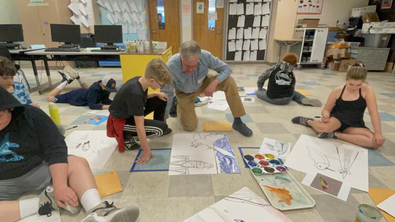 A White man kneels on the floor to help a group of students review their artwork.