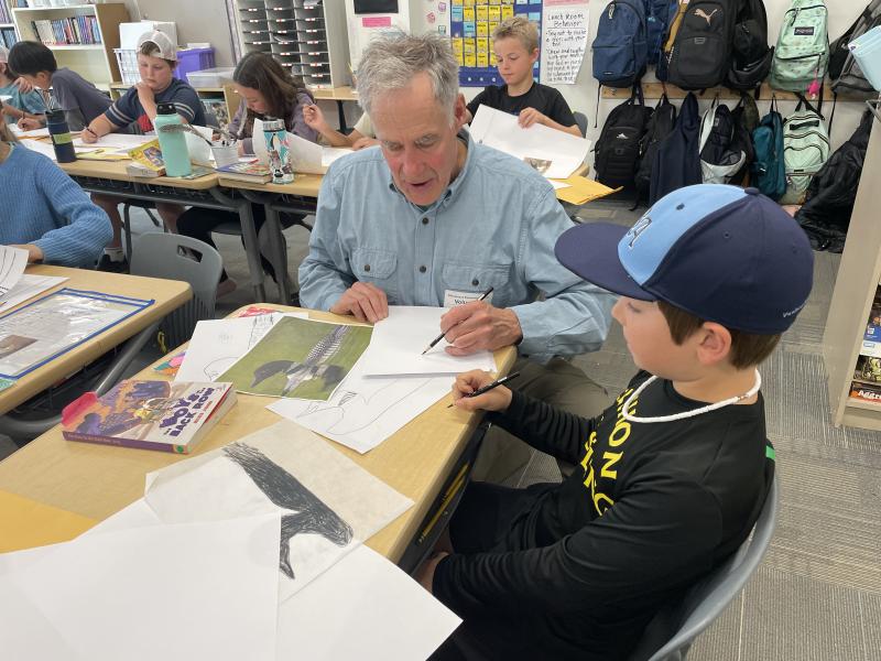 A White man sits in a classroom full of students, helping a young White boy with his artwork.