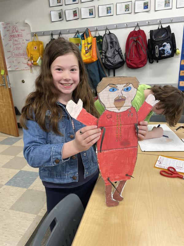 A young White girl with long, brown hair holds a cardboard puppet.