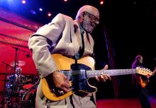 Man in a beige suit plays the electric guitar with a man on drums and another man on guitar in the background