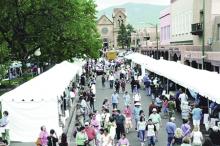 Crowds of people walking down the street and looking into various white tents lined up along the curb; an old church in the background.