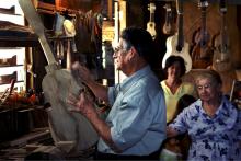 A man works on building a stringed instrument with others behind him watching.