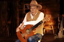 A man in a brown cowboy hat and best sits on a stool, holding a guitar.