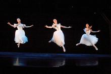 Three female ballerinas jumping onstage in white outfits as they perform. 