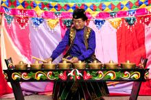 A man in a purple shirt and gold and black vest sits behind a musical instrument made up of eight small gongs.