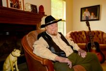 A man wearing a cowboy hat sits in a chair with a guitar next to him.