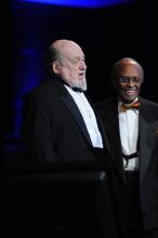 A man in a tuxedo speaks from a podium while another man in a brightly colored tie looks on smiling